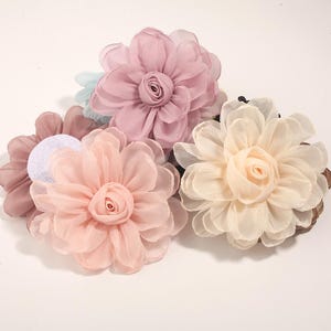 Może przedstawiać: Kolekcja akcesoriów do włosów z kwiatami z tkaniny w różnych pastelowych kolorach. Kwiaty są w odcieniach różu, brzoskwini i kremu, z delikatnymi detalami płatków. Odpowiednie na wesela, imprezy lub na co dzień.