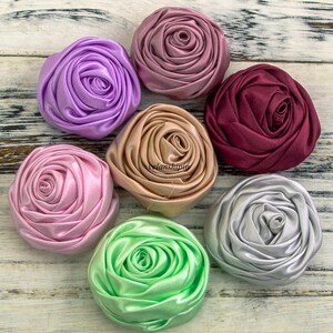 May include: Seven fabric flower embellishments in various colors, including lavender, pink, beige, burgundy, silver, and green. The flowers are made of satin ribbon and have a rosette design.