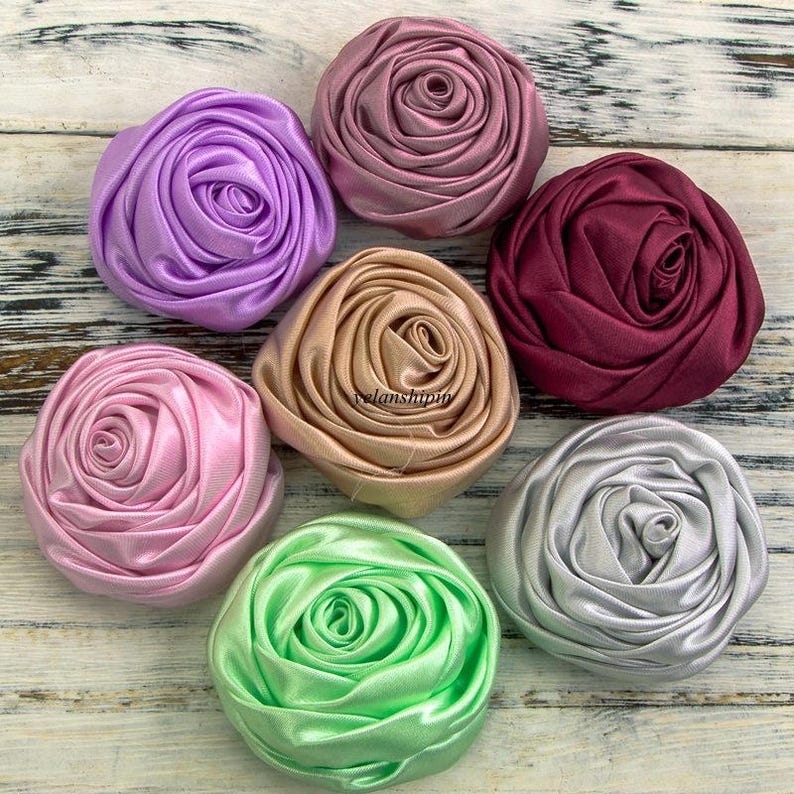 May include: Seven fabric flower embellishments in various colors, including lavender, pink, beige, burgundy, silver, and green. The flowers are made of satin ribbon and have a rosette design.
