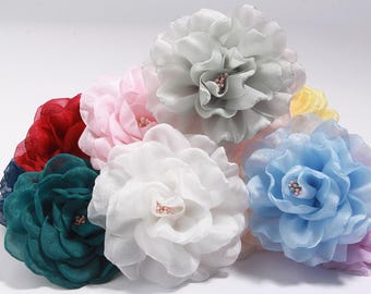 May include: A collection of fabric flower embellishments in various colors, including white, pink, blue, and green. Each flower has multiple layers of petals and a small cluster of beads in the center. The flowers are arranged on a white surface.