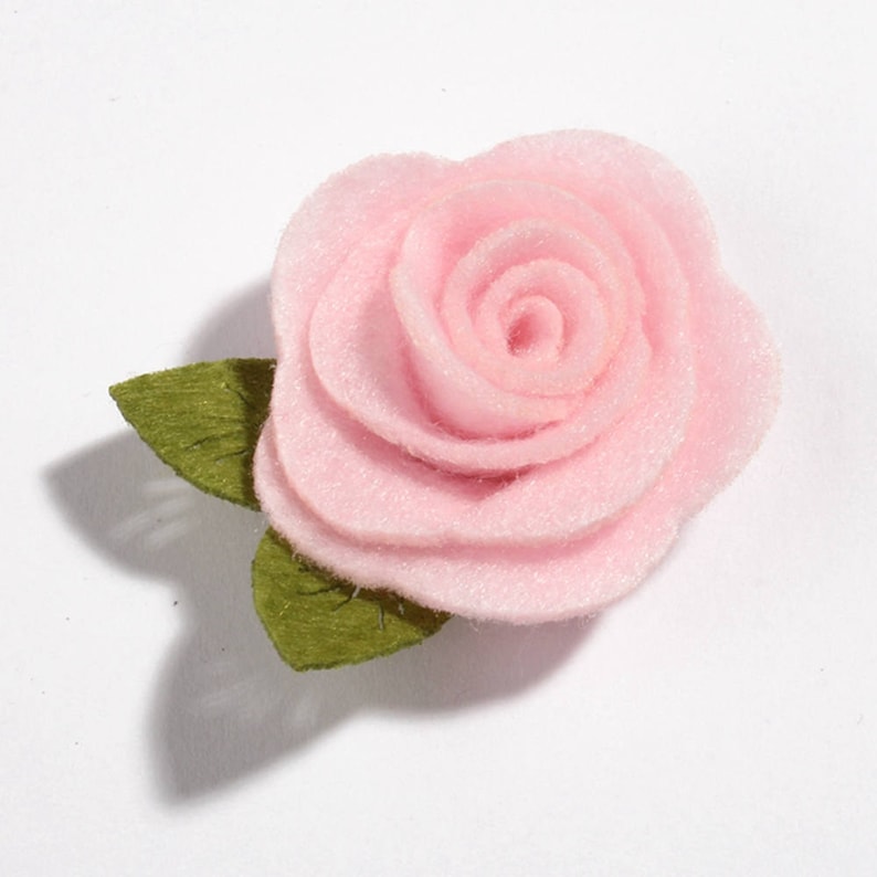 K&ouml;nnte beinhalten: Eine hellrosa Filzrose mit gr&uuml;nen Filzbl&auml;ttern. Die Rose hat mehrere Schichten von Bl&uuml;tenbl&auml;ttern, wodurch ein realistisches Aussehen entsteht. Die Bl&auml;tter sind einfach und flach gestaltet. Geeignet f&uuml;r verschiedene Bastelprojekte.