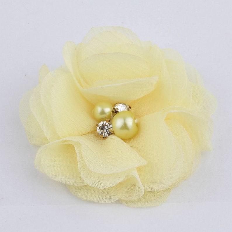 May include: A pale yellow fabric flower with a layered design. The center of the flower is adorned with three pearl-like beads and a clear rhinestone.