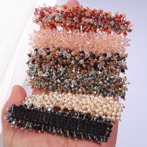 Può includere: Un set di sei fermagli per capelli, ognuno decorato con perline di cristallo di un colore diverso. I fermagli sono disposti in pila, con il fermaglio superiore di colore rosso, seguito da rosa, nero, verde, bianco e nero.