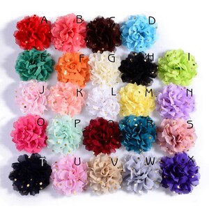 Op de afbeelding: Een verzameling van vierentwintig kleurrijke stoffen bloemversieringen. Elke bloem heeft een andere kleur, waaronder rood, roze, blauw, groen en geel. De bloemen hebben een gelaagd bloemblaadjesontwerp en zijn versierd met goudkleurige pailletten.