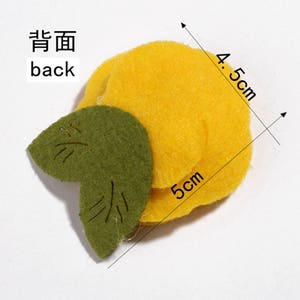 K&ouml;nnte beinhalten: Ein Bastelartikel aus Filz, der einer Zitrone mit einem gr&uuml;nen Blatt &auml;hnelt. Die Zitrone ist gelb und misst 5 cm x 4,5 cm. Das Blatt ist gr&uuml;n mit gestickten Details. Das Wort "back" ist auf Englisch und Chinesisch geschrieben.