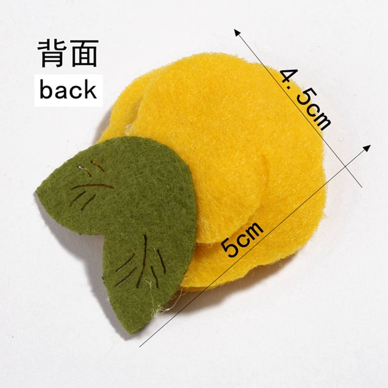 K&ouml;nnte beinhalten: Ein Bastelartikel aus Filz, der einer Zitrone mit einem gr&uuml;nen Blatt &auml;hnelt. Die Zitrone ist gelb und misst 5 cm x 4,5 cm. Das Blatt ist gr&uuml;n mit gestickten Details. Das Wort "back" ist auf Englisch und Chinesisch geschrieben.