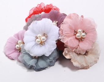 Puede incluir: Una colección de adornos florales de tela en varios colores, incluyendo rosa, blanco, rojo y gris. Cada flor presenta un grupo de perlas de imitación y pequeños diamantes de imitación en el centro, con delicados bordes de encaje.