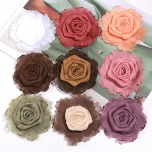May include: Nine fabric rose embellishments in various colors, including white, red, peach, brown, pink, green, tan, and purple. Each rose has a layered petal design and a ruffled edge, suitable for crafts or decorations.