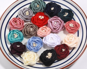 Puede incluir: Una colección de adornos florales de tela colorida en varios tonos de rosa, rojo, azul, verde y blanco, dispuestos sobre un plato blanco con rayas azules. Cada flor tiene un pequeño centro de cuentas blancas.