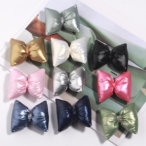 Könnte beinhalten: Eine Auswahl an Satin-Schleifen-Haarschmuck in verschiedenen Farben, darunter Gold, Silber, Rosa, Schwarz und Blau. Die Schleifen haben ein puffiges Design und sind auf einer gemusterten Oberfläche angeordnet.