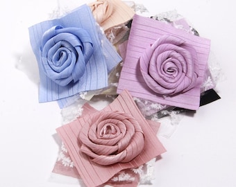 Puede incluir: Una colección de adornos florales de tela hechos a mano en varios colores pastel. Cada flor tiene forma de rosa, unida a un cuadrado de tela a rayas y encaje. Los colores incluyen azul, rosa y lavanda.