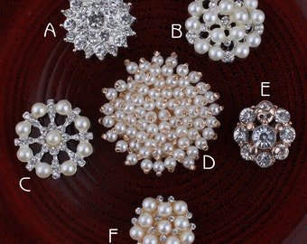 May include: Six different types of embellishments for crafts and sewing projects. The embellishments are round and feature a variety of designs, including pearl clusters, rhinestones, and a snowflake design. The embellishments are arranged on a red surface.