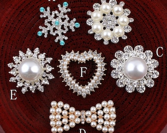 May include: Assortment of six pearl and rhinestone embellishments in various shapes, including a snowflake, a heart, a circle, and a bow. The embellishments are silver-toned and feature white pearls and clear rhinestones.