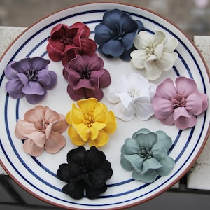 Puede incluir: Una colección de flores artificiales coloridas en varios tonos, incluyendo rojo, morado, amarillo y azul, dispuestas sobre un plato blanco con rayas azules. Las flores tienen múltiples pétalos y probablemente están hechas de tela o material similar.