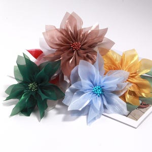 May include: A collection of five artificial flowers in various colors: brown, green, blue, and gold. Each flower has delicate, translucent petals and a beaded center. The flowers are arranged on a white surface, suggesting a craft or decorative theme.