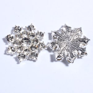 Könnte beinhalten: Zwei silberne Schneeflocken-Verzierungen. Eine ist ein Cluster aus klaren Strasssteinen, die andere ist eine massive Schneeflocke aus Metall.