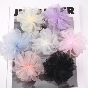 May include: A collection of seven sheer fabric flower embellishments in various pastel colors, including white, light blue, lavender, and pink. The flowers are arranged on a magazine page, with the word "JENNIFER" visible.