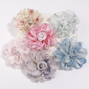 Może przedstawiać: Sześć materiałowych kwiatów w pastelowych odcieniach. Kwiaty mają warstwowe płatki i centralny pączek. Kolory obejmują różowy, niebieski, lawendowy i kremowy.