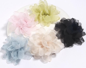 Puede incluir: Cinco adornos florales de tela en colores pastel: rosa claro, amarillo pálido, crema, azul claro y negro. Cada flor tiene múltiples capas de pétalos transparentes, creando una apariencia delicada y texturizada. Las flores están dispuestas sobre un plato blanco.