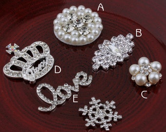 May include: Assortment of silver and white rhinestone embellishments, including a crown, a snowflake, a word "love", a flower with pearls, and a circle of pearls.