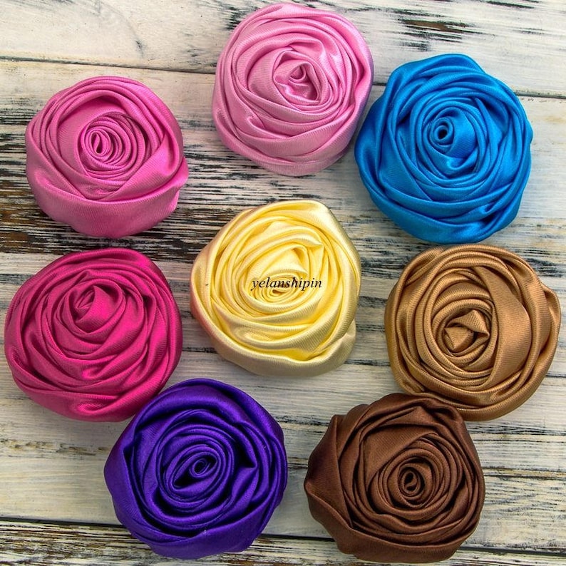 May include: Assortment of eight handmade fabric rosettes in various colors, including pink, blue, yellow, fuchsia, purple, brown, and gold. The rosettes are made of satin fabric and have a layered, ruffled appearance.