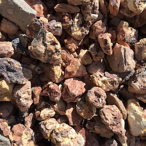 May include: A close-up view of a pile of natural, rough-cut rocks. The rocks vary in size and shape, with a mix of reddish-brown, tan, and gray hues. Some rocks have dark, almost black, mineral deposits. The overall texture is coarse and natural.