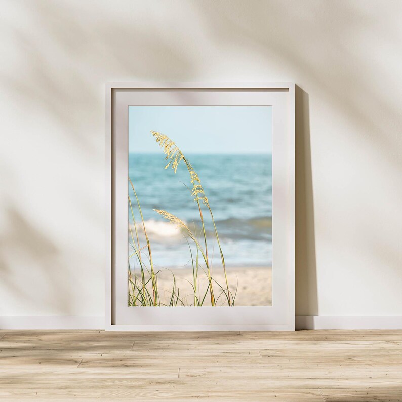 May include: A framed print featuring a coastal scene. The artwork depicts tall, golden sea oats in the foreground, with a sandy beach and the ocean in the background. The sky is a soft blue, and the frame is white.