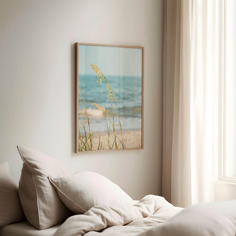 May include: A framed print featuring a beach scene with tall, wispy grasses in the foreground. The artwork showcases a sandy beach, the ocean, and a light blue sky. The frame is a light brown color, and the overall aesthetic is calming and natural.