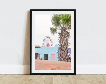 May include: A framed wall art print featuring a light blue building with a pink window, a tall palm tree, and a colorful Ferris wheel in the background under a bright sky. The print is enclosed in a simple black frame and displayed on a wooden floor.