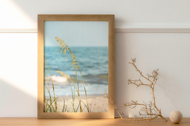 May include: A framed print featuring a coastal scene with sea oats and ocean waves. The wooden frame surrounds the image of a beach with tall grasses and a serene blue sea. A decorative branch and sphere are on the table.