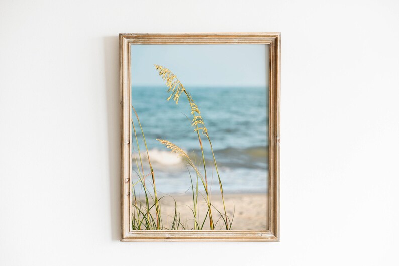 May include: Framed beach scene print featuring tall, golden sea oats in the foreground, with a sandy beach and the ocean in the background. The water is a light blue, and the frame is a light brown color.