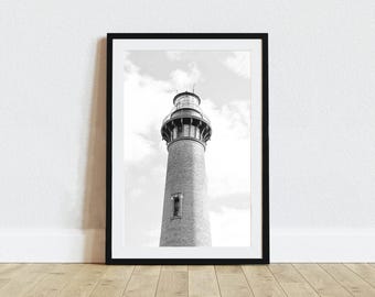 May include: A black and white framed wall art print featuring a tall lighthouse with a brick texture, a lantern room at the top, and a single window on the side, set against a cloudy sky. The print is enclosed in a simple black frame.