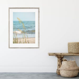 May include: Framed beach scene print featuring a coastal landscape with sea oats in the foreground. The artwork depicts a sandy beach, ocean waves, and a light blue sky. The frame is a light gray color.