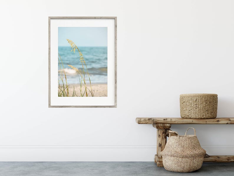 May include: Framed beach scene print featuring a coastal landscape with sea oats in the foreground. The artwork depicts a sandy beach, ocean waves, and a light blue sky. The frame is a light gray color.