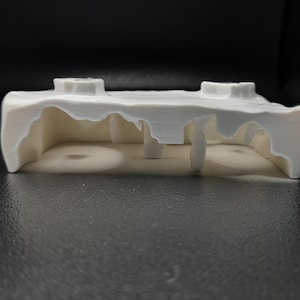 Peut inclure: Objet blanc imprimé en 3D avec un design architectural unique. La structure présente une base rectangulaire avec un dessus irrégulier et texturé et plusieurs supports intérieurs. Deux protubérances cylindriques sont visibles sur la surface supérieure.