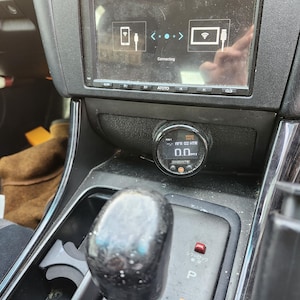 May include: A black car interior with a black and silver gauge that reads "0.0" and the text "PSI x 0.021 BAR". The gauge is mounted on the dashboard next to a black gear shift lever.