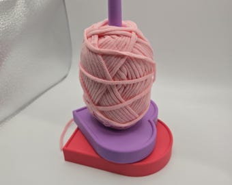 May include: A pink yarn ball sits atop a three-tiered, heart-shaped yarn holder. The holder is composed of a red base, a purple middle layer, and a pink top. A pink peg rises from the yarn ball.