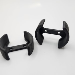 May include: Two black, 3D-printed, curved brackets. Each bracket has a horizontal bar with two holes, and two curved arms. The arms have vertical ridges. The brackets are on a white surface.
