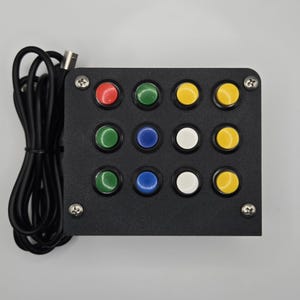 Peut inclure: Un panneau de commande noir avec douze boutons multicolores. Les boutons sont rouges, verts, jaunes, bleus et blancs. Un câble noir avec un connecteur est attaché au panneau.