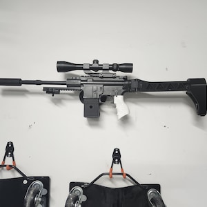 May include: A black rifle with a scope mounted on top. The rifle has a white plastic grip and a black stock. The rifle is mounted on a white wall.