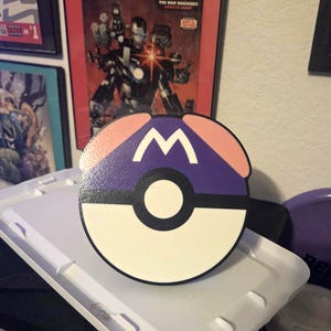 May include: A round, wooden decorative item featuring a design inspired by a Pokeball and the letter "M". The item is primarily white, purple, and pink, with a black stripe and a white "M" in the center. The item is sitting on a white tray.