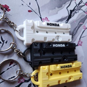 Pode incluir: Três chaveiros com um design de bloco de motor Honda. Os chaveiros são branco, preto e amarelo.