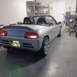 Könnte beinhalten: Ein silbernes Cabriolet mit einem grünen Nummernschild, auf dem "SMLPP" steht, und einem schwarzen Heckspoiler mit dem Wort "TAVZI" darauf. Das Auto steht in einer Garage mit einem Motorrad im Hintergrund.