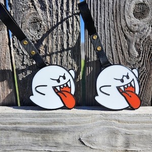 May include: Two white ghost-shaped luggage tags with black outlines, featuring a red tongue and a black strap with gold rivets. The tags are displayed against a weathered wooden fence.