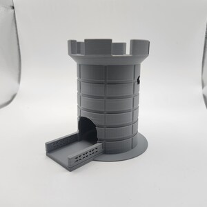 May include: A gray 3D-printed dice tower, resembling a castle tower. The tower has a cylindrical body with horizontal bands, a crenelated top, and a ramp for dice to exit. The base is circular.