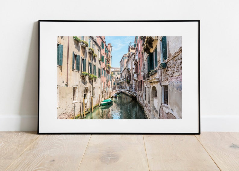 K&ouml;nnte beinhalten: Ein schmaler Kanal in Venedig, Italien, mit farbenfrohen Geb&auml;uden an den Seiten. Ein kleines gr&uuml;nes Boot befindet sich im Wasser, und eine Br&uuml;cke &uuml;berquert den Kanal in der Ferne.
