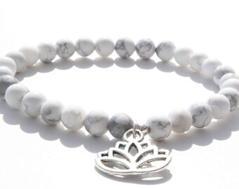 May include: A stretch bracelet composed of round white and grey marbled beads. The bracelet features a silver-toned metal charm shaped like a stylized lotus flower hanging from the center of the bead strand, creating a circular accessory design.