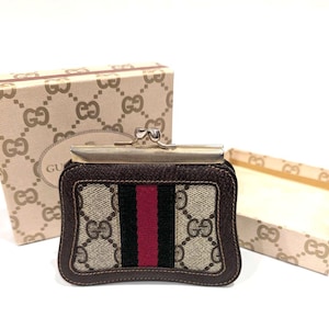 Puede incluir: Un monedero marrón y beige con cierre metálico, con un diseño de rayas rojas y negras. El monedero va acompañado de una caja a juego con un patrón de logotipo repetido. La palabra "GUCCI" es visible en la caja.