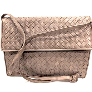 May include: A taupe-colored, woven leather shoulder bag with a flap closure. The bag features a long, adjustable strap and a knotted detail at the end. The bag's design is a repeating pattern of woven leather.