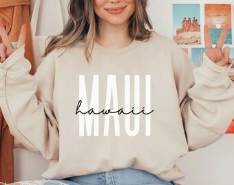 Puede incluir: Sudadera beige con la palabra "MAUI" en fuente vertical y en negrita y "hawaii" en cursiva. La sudadera es de color neutro y tiene mangas largas. El tema general es viajes y vacaciones.